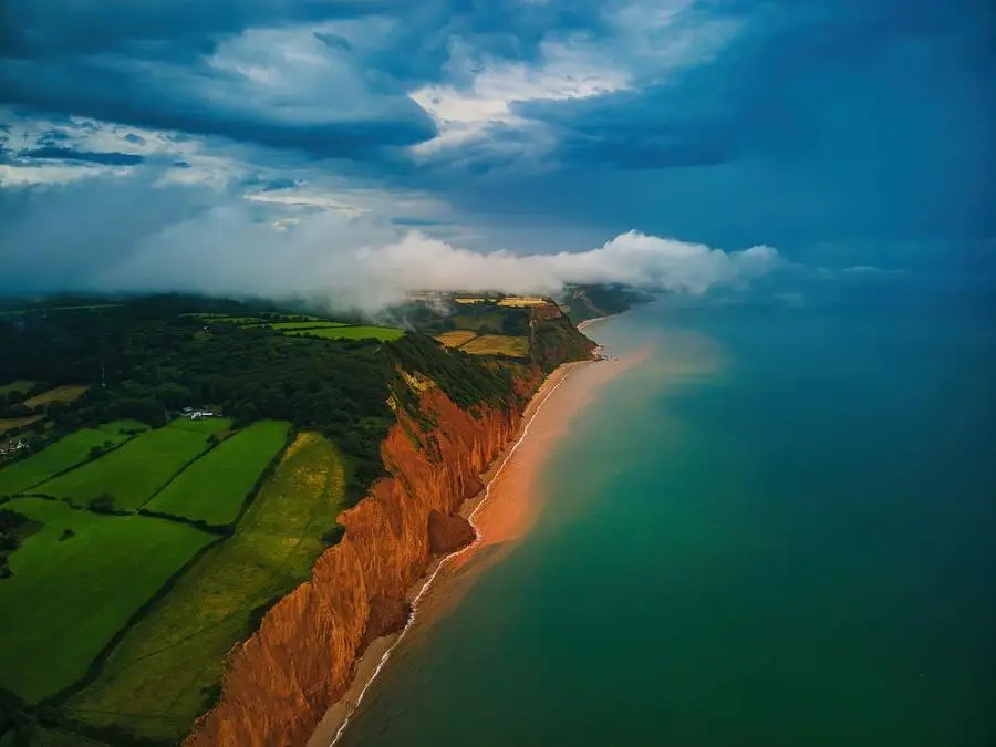 Aerial view of green fields above a red cliff face bordered by the sea.