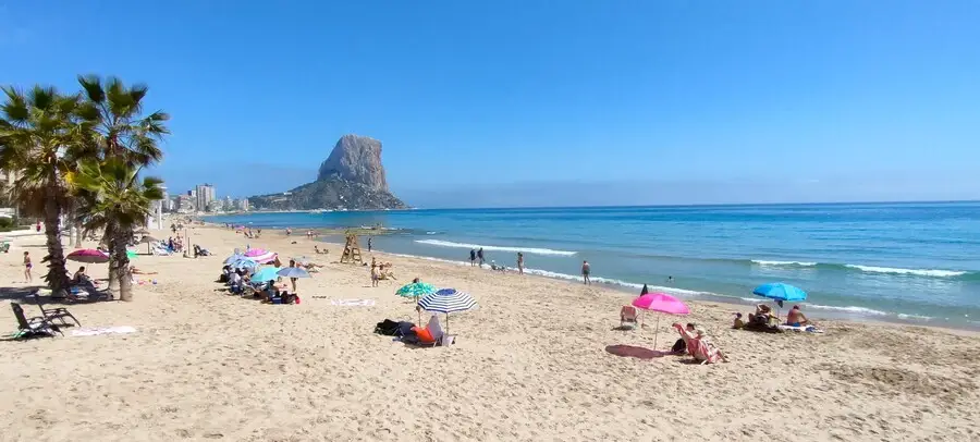 Tourists sitting on sandy Arenal-Bol Beach with 'Calpe rock' in the distance.