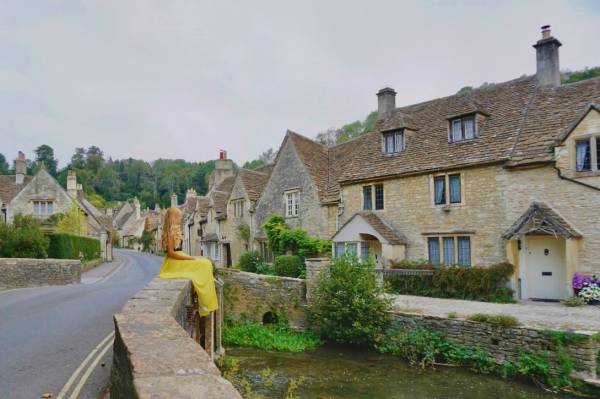 Woman sitting beside old stone river bridge & village houses in Castle Coombe, Cotswolds.