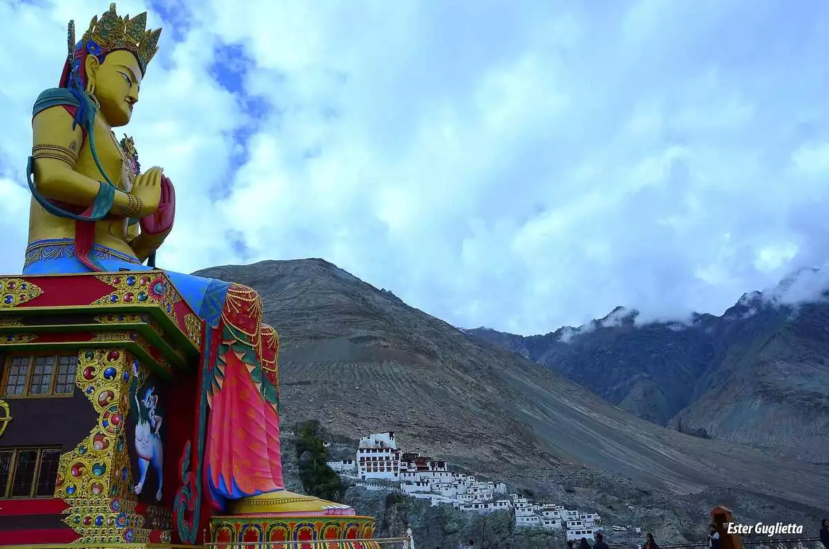 Gran estatua dorada de Buda sentada en las montañas de Ladakh con un templo al fondo.