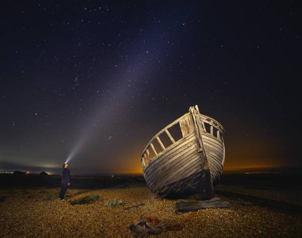 Shining torch on starlit sky, boat on shingle beach in Dungeness, Kent.