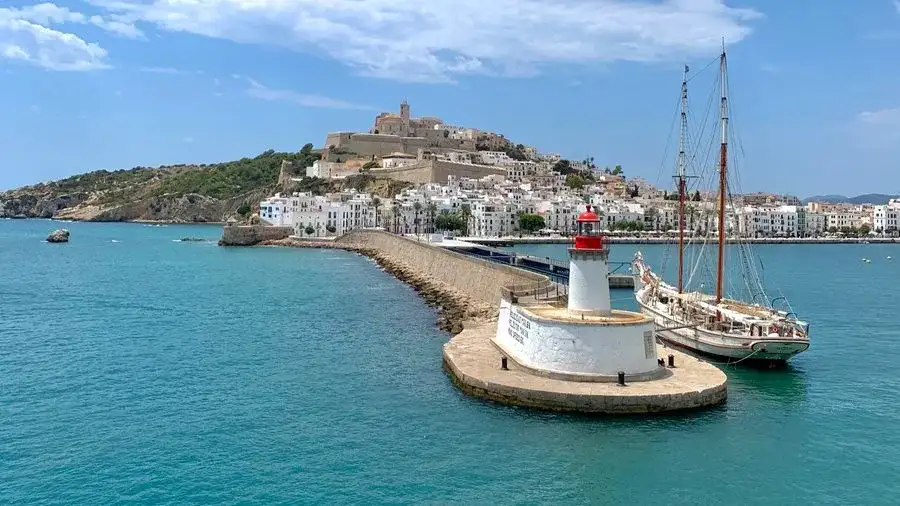 Tall mast sailing boat in the sea with walled Ibiza old town in the distance.