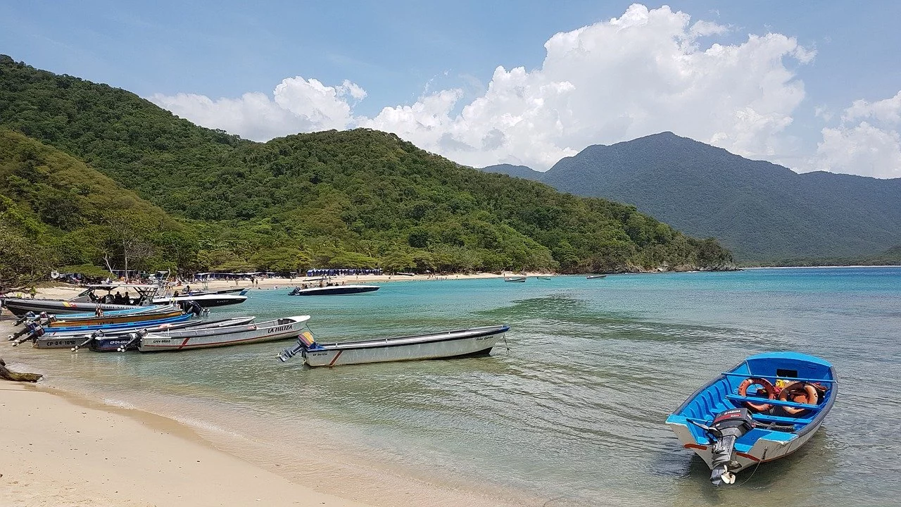 Bahia con arena, colina arbolado y mar Caribe con pequeñas embarcaciones caribeñas.