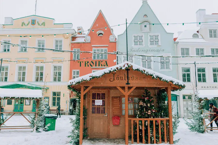 Snowy scene of wood Christamas hut in front of traditional Tallinn buildings.
